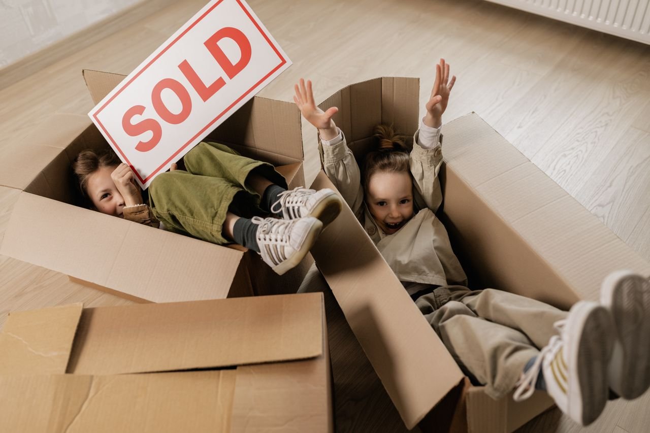gallery-02 Two happy children playing with cardboard boxes indoors, celebrating moving to a new home.